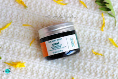 A high-angle shot of The Body Shop Vitamin C Glow Boosting Moisturiser jar, featuring its dark green glass, silver lid, and white and orange label, resting on a textured white surface with scattered yellow petals.