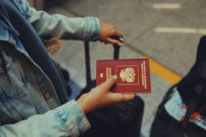 A close-up shot of a person's hands holding a red Russian passport while gripping the handle of a black rolling suitcase.