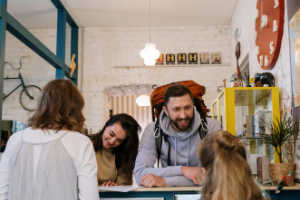 A group of four travelers, including a man with a large backpack, are smiling and talking to a person at a reception desk, likely checking into a hotel or hostel.