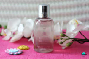 
A clear bottle of Molton Brown Delicious Rhubarb & Rose bath and shower gel is positioned in the center of the frame. The bottle is placed on a vibrant pink surface. Surrounding the bottle are a white orchid and two small, decorative flowers, one yellow and one pink. The background is a striped pattern. Featured on everything enchanting blog.