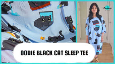 A split-panel image promoting a review of the Oodie Black Cat Sleep Tee. The left panel is a close-up of the light blue fabric with the playful black cat print, showing a kitten on a laptop and another lying down. The right panel shows uk fashion blogger Anamika  standing and modeling the oversized sleep tee, highlighting its generous fit. A large white bubble with black text reads "OODIE BLACK CAT SLEEP TEE".