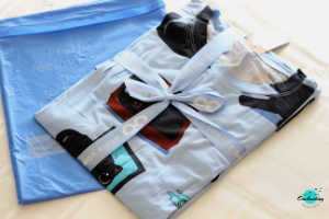 A light blue Oodie Black Cat Sleep Tee featuring an all-over print of playful black cats is folded neatly and tied with a matching blue ribbon, suggesting gift-ready packaging. The folded tee rests beside a matching light blue pouch or storage bag, showcasing the attention to detail in the product presentation. Everything Enchanting blog logo is featured at the right corner of the image. 