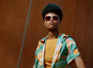 A person with short, dark curly hair and red-tinted sunglasses stands against an orange-brown background. They are wearing a bright, patterned short-sleeve button-down shirt over a fitted yellow/orange ribbed top.