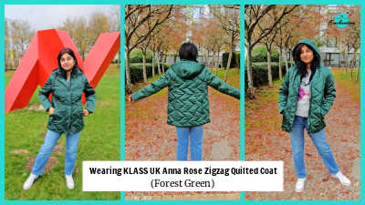 A woman stands on an outdoor path covered with red and brown autumn leaves. She is wearing a dark forest green hooded quilted coat with a diamond zig-zag pattern, layered over a light grey argyle knit jumper. The outfit is completed with straight-leg blue jeans and white sneakers.