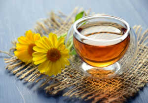 A close-up, slightly high-angle shot of a clear, double-walled glass cup filled with amber-colored herbal tea. The cup sits on a rustic, frayed burlap coaster atop a blue wooden surface. Two bright yellow daisy-like flowers with orange centers rest next to the cup, adding a natural and calming touch to the scene.
