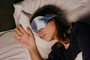 A close-up, top-down shot of a person sleeping peacefully on a white pillow with their head tilted slightly to the side. They are wearing a silky, light-blue sleep mask over their eyes and a dark navy blue long-sleeved top. One hand is resting open on the pillow near their face.