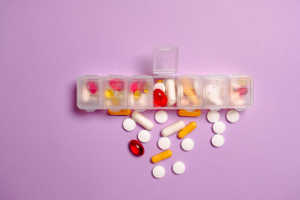 A top-down shot of an open, clear plastic seven-day pill organizer resting on a solid purple background. The compartments are filled with a variety of colorful vitamins and supplements, including red softgels, yellow capsules, and white tablets. Additional loose pills in various shapes and colors are scattered directly below the organizer on the purple surface.
