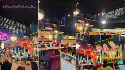 The interior of a high-end Mediterranean restaurant featuring deep plum velvet seating and marble-topped tables set with red plates and stemless glassware. The double-height space is illuminated by ornate, tiered crystal and gold chandeliers hanging from a dark industrial ceiling. A mezzanine level overlooks the main floor, partially obscured by an intricate dark laser-cut geometric screen. Lush botanical installations, including cascading pink wisteria and a vibrant red maple-style tree, create organic partitions throughout the dining area. In the background, a gold-textured geometric bar front sits beneath a large black awning that marks the open kitchen.