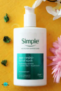 Flat lay of a 240ml bottle of Simple Age Resist+ Facial Wash with its signature white pump and teal label, styled on a vibrant yellow textured background with pink flowers and dried green leaves.