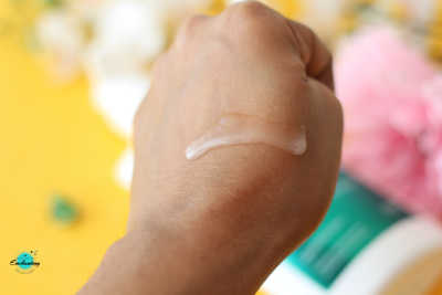 Close-up of the clear, translucent gel texture of Simple Age Resist+ Facial Wash swatched on the back of a hand, showcasing its smooth and lightweight consistency against a bright yellow background with pink floral accents.