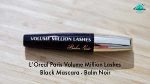 A tube of L'Oréal Paris Volume Million Lashes Balm Noir mascara in black, lying horizontally on a light-colored wooden surface. The mascara tube features a sleek black body with gold lettering and a metallic gold cap. Text at the bottom of the image reads "L'Oreal Paris Volume Million Lashes Black Mascara - Balm Noir."