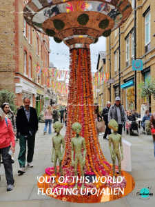 A vertical photograph of a large, outdoor art installation on a busy brick-lined street. The installation features a large, metallic, saucer-shaped UFO structure elevated above the ground. Cascading down from the center of the saucer to a circular orange base on the pavement are long strands of orange and red flowers, simulating a tractor beam. Standing in front of the floral beam are three small, green alien figures. Pedestrians walk on either side of the display, and festive, colorful ribbons stretch across the street in the background. White text at the bottom reads, "'OUT OF THIS WORLD' FLOATING UFO/ALIEN".