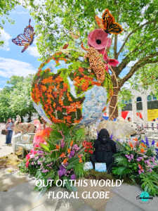 A vertical photograph of a large, outdoor floral installation representing a globe. The globe structure is crafted from green moss and orange, red, and white flowers to depict the continents and oceans. Surrounding the base of the globe are lush tropical plants, purple flowers, and a dark gorilla statue sitting on the right. Atop the globe, a large pink flower blossom and an orange monarch butterfly model are attached, with another colorful butterfly model seemingly floating in the bright blue sky to the upper left. Green trees frame the background. White text at the bottom reads, "'OUT OF THIS WORLD' FLORAL GLOBE".