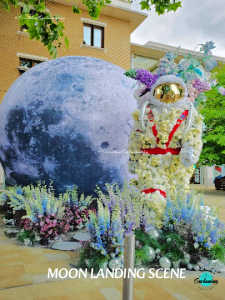 A vertical photograph of a themed outdoor art display. To the left is a large, spherical representation of the moon with textured craters. To the right stands a life-sized astronaut figure, whose suit is intricately covered in cream-colored flowers with a bright gold helmet visor. The astronaut is surrounded by a base of blue, purple, and green floral accents, mimicking a lunar landscape. In the background, a brick building and green trees are visible. White text at the bottom reads, "MOON LANDING SCENE".