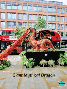 A vertical photograph of a large, outdoor floral installation featuring a mythical dragon sculpture. The dragon is primarily reddish-orange, with dark, textured wings and a long tail, and it appears to be exhaling a stream of vibrant orange and red flowers. The sculpture is mounted on a black rectangular base surrounded by green ferns and potted plants. In the background, a bright red double-decker bus is parked on a city street, partially obscured by the dragon display, in front of a modern multi-story brick building with many windows. White text at the bottom reads, "Giant Mythical Dragon".