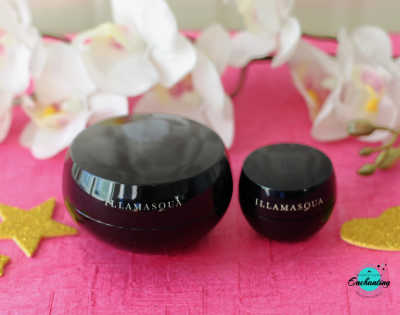 A product photograph of two glossy black, spherical jars of Illamasqua Hydra Veil primer in different sizes—a large 30 ml jar on the left and a small 7 ml travel jar on the right. Both jars feature silver "ILLAMASQUA" branding and are placed on a vibrant pink textured surface. The background is softly blurred with white orchid flowers, and a small blue "Enchanting" logo is visible in the bottom right corner.