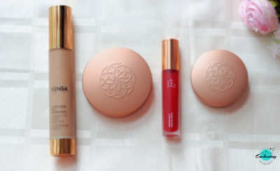 The image displays a collection of YENSA beauty products arranged on a light surface, including a foundation bottle, two closed compacts with rose-gold lids embossed with a geometric logo, and a bright pink lip oil, with pink artificial flowers in the upper right corner and an "Enchanting" logo in the lower right corner.
