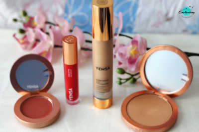 The image features a collection of YENSA makeup products on a white surface, including an open cream blush compact, a vibrant pink lip oil, a tall bottle of foundation, and an open powder foundation compact, all displayed in front of a branch of pink orchids with an "Enchanting" logo in the upper right corner.