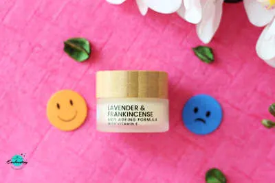 A small jar of The Edinburgh Natural Skincare Co. Lavender & Frankincense body butter centered on a vibrant pink background, flanked by a yellow happy face icon on the left and a blue sad face icon on the right to represent pros and cons. Review photo featured on Everything Enchanting blog.