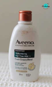 A white bottle of Aveeno Scalp Soothing Haircare Volumising Shampoo for Fine Hair, featuring the Fresh Greens Blend formula, resting on a white textured surface with the Everything Enchanting logo in the top-right corner.