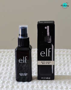 A black spray bottle of e.l.f. Matte Magic Mist & Set standing beside its matching black cardboard box on a white textured surface, with the Everything Enchanting logo in the top-right corner