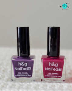 Two bottles of Health & Glow (h&g) Nail'ed It Nail Enamel—one in a dark violet shade on the left and a bright pink shade on the right—standing side-by-side on a white textured knitted surface, with the Everything Enchanting logo in the top-right corner.