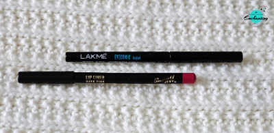 A black Lakmé Eyeconic Kajal pencil and a Barry M Rose Pink Lip Liner pencil lying parallel on a white textured knitted background with the Everything Enchanting logo in the top-right corner.