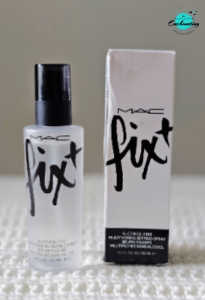 A frosted clear bottle of MAC Prep + Prime Fix+ setting spray standing next to its white and black cardboard box on a textured white surface, with the Everything Enchanting logo in the top-right corner.