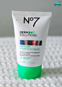 A white tube of No7 Derm Solutions 100-Hour Hydration Cream for dry and sensitive skin, featuring a bright green base cap, standing on a textured white surface with the Everything Enchanting logo in the top-right corner.