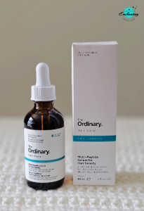 An amber glass dropper bottle of The Ordinary Multi-Peptide Serum for Hair Density standing beside its white cardboard packaging box on a white textured surface, with the Everything Enchanting logo featured in the top-right corner.