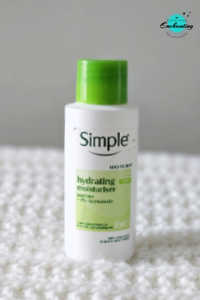 A travel-size white bottle of Simple Kind to Skin Hydrating Light Moisturiser topped with a bright green cap, standing vertically on a white textured knitted background with the Everything Enchanting logo in the top-right corner.