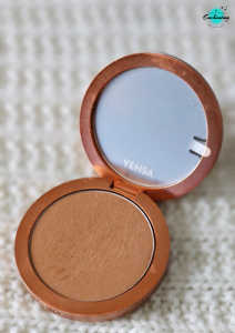 An open circular bronze compact of Yensa Super Silk Powder Foundation resting on a white textured knitted surface, revealing the pressed powder in Medium Warm shade and a round inner mirror with the Yensa logo.